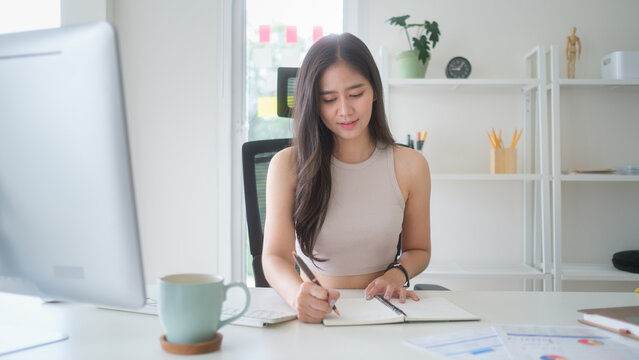Young businesswoman writing notes in a notebook while working at her desk in a bright modern office with laptop and coffee mug.