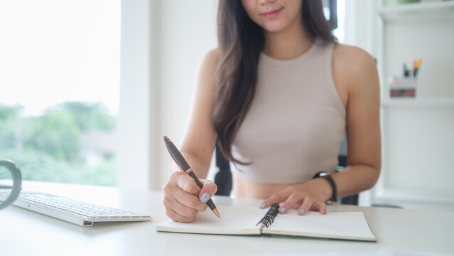 Young businesswoman writing notes in a notebook while working at her desk in a bright modern office with laptop and coffee mug.