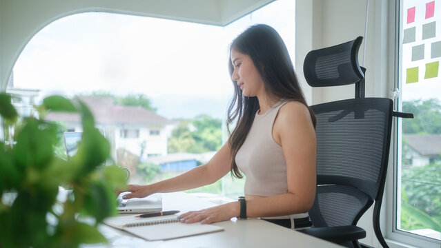 Smiling young businesswoman working on a laptop at her desk in a bright home office with large windows and natural daylight.