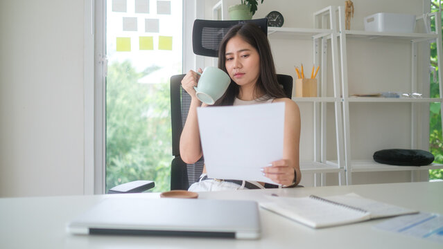 Smiling young businesswoman holding a cup of coffee while reading a document in a bright and modern office.