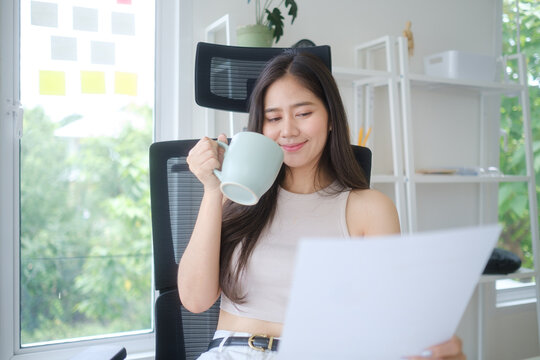Smiling young businesswoman holding a cup of coffee while reading a document in a bright and modern office.