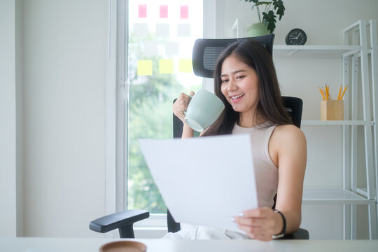 Smiling young businesswoman holding a cup of coffee while reading a document in a bright and modern office.