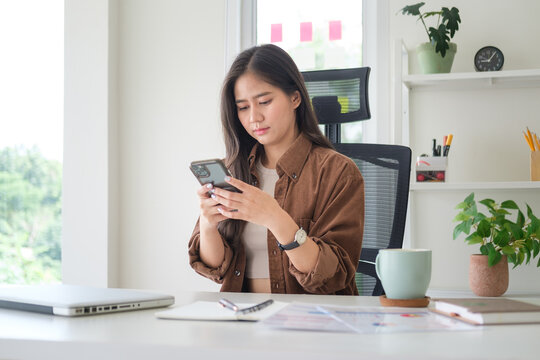 Young businesswoman checking messages on her smartphone while sitting at a desk with laptop, documents, and coffee in a bright modern office.