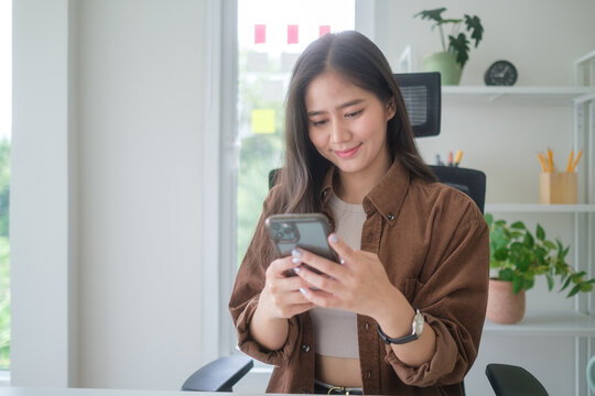 Young businesswoman checking messages on her smartphone while sitting at a desk with laptop.