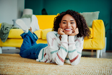 Bright happy young woman with curly hair lies on a rug in a colorful striped sweater at home on a...