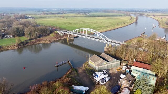 A white arched bridge spans a calm river curving through open fields, with boats, sheds, and small buildings along the shore, forming a clear and peaceful rural landscape.