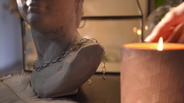Woman putting a stylish silver chain necklace on a plaster bust of a head on a dressing table. The scene is illuminated by a warm candle