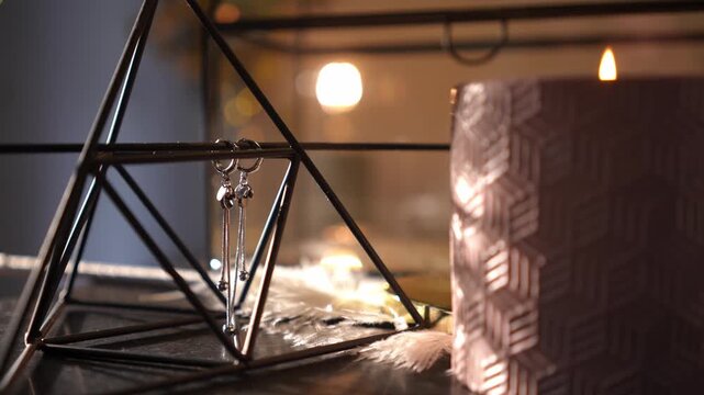 Close-up of a cozy, elegant still life. A burning candle's warm light flickers, creating moving shadows over silver earrings and decor