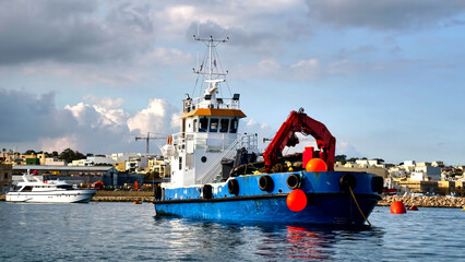 Marsaxlokk, Malta &ndash; Workboat with crane arm, buoys and fishing nets in front of townscape and cloudy sky