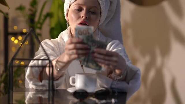 Glamorous young woman in a bathrobe and towel on her head counting a stack of hundred-dollar bills while sitting at a table in the morning