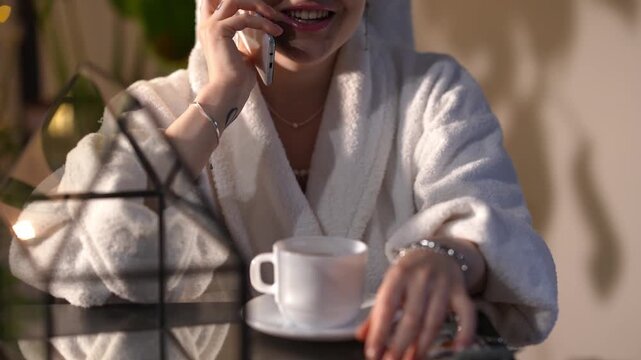 Young woman in a white bathrobe relaxing at a spa. She is sitting at a table, drinking coffee, and pleasantly talking on a smartphone