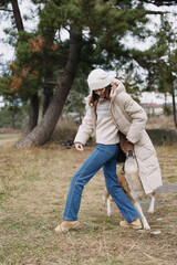 Person walking dog in an outdoor park, woman in a long coat and hat, jeans and boots, casual fashion pose amidst autumn scenery and leash, capturing companionship