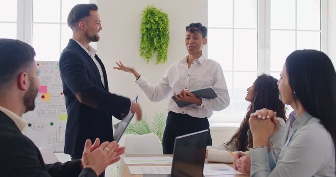 Young female leader introducing the best employee of month, exchanging high five gesture, celebrate joint success during meeting led by boss standing in front of staff members, presenting newcomer