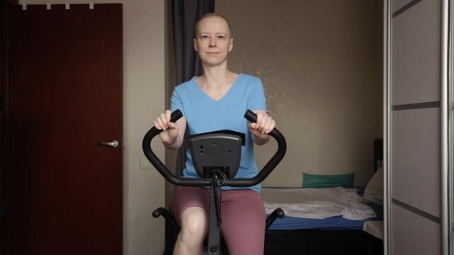 Female cancer patient doing cardio training on home exercise bike, safe workout during immunosuppression, rehabilitation program at home, healthy habits and mental health support