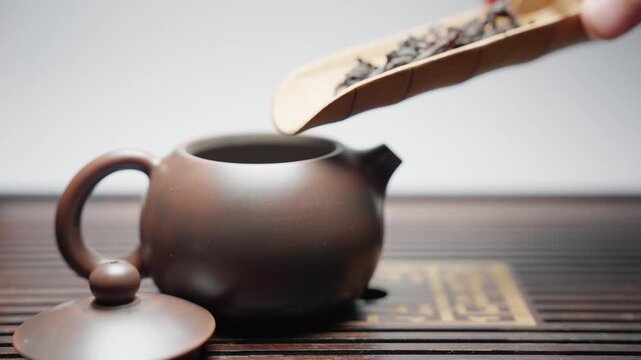 Side view close-up in slow motion. Dry tea leaves are poured from a chahe into an empty brown clay teapot on a traditional tea board, completing a mindful preparation ritual.