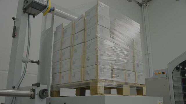 Pallet of packaged boxes being wrapped in plastic film by automated machinery in a warehouse setting, showcasing the continuous wrapping process and equipment operation, hdr video with neutral color
