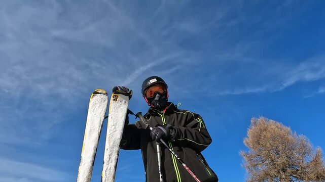 Portrait of skier in helmet goggles and face mask standing with skis against blue sky in Folgaria Italy, winter gear fashion