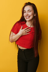 Obraz premium Young woman with long hair smiling and winking with hand on chest wearing red shirt and black pants standing against yellow background. Positive emotion and casual style portrait.