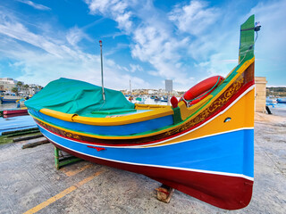 Colorfully painted Luzzu fishing boat on land with tarp, traditional ornaments, blue sky in Marsaxlokk