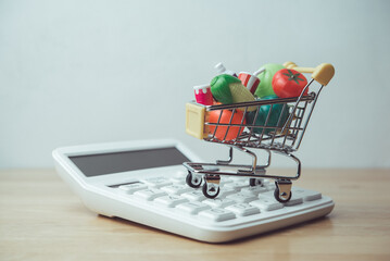 Vegetables, fruits, drinks, food material in supermarket trolley and calculator on wooden table. Rising cost of living, food price increase, inflation crisis, home finance and spending money concept.