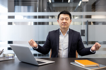 Fototapeta premium Asian businessman in a suit finding inner peace and mindful focus through meditation at his modern office workspace, promoting calm and well-being during a busy workday