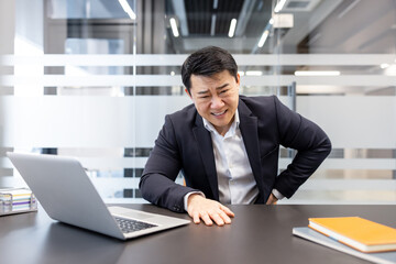 Fototapeta premium Asian businessman sitting at his desk, experiencing severe lower back pain and discomfort while working on a laptop in a modern office, highlighting work-related health issues