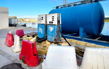 Modern blue diesel tank system with old rusty fuel pumps and barriers in foreground at Marsaxlokk harbor, Malta.
