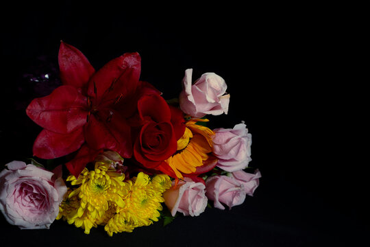 Elegant floral bouquet featuring a deep red lily, a classic red rose, and bright yellow chrysanthemums against a solid black background. Moody, high-contrast still life photography with rich colors