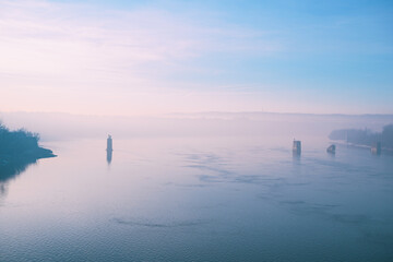 Foggy winter scene of the Danube river at dawn, featuring still water, pale sky, bridge remains, and peaceful atmospheric mood with copy space.