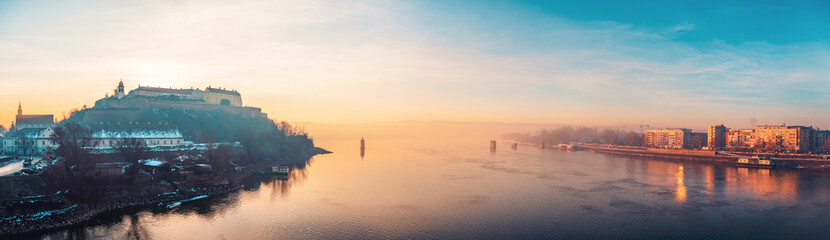 Wide panorama of the Danube river at dawn, showing Petrovaradin Fortress and Novi Sad waterfront, misty winter morning, tranquil water, and pastel sky.
