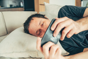 Unhappy middle-aged man resting on sofa with tired eyes, doomscrolling on mobile phone, expressing depression, exhaustion, or emotional distress in a realistic domestic setting.