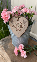 Rustic galvanized metal bucket overflowing with vibrant pink carnation flowers featuring a wooden heart tag engraved with happy mothers day text suspended by jute twine on a weathered timber surface.