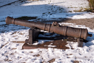 Old rusty cannon displayed outdoors on a wooden stand in winter. Historical artillery weapon covered by light snow in a park or fortress area.