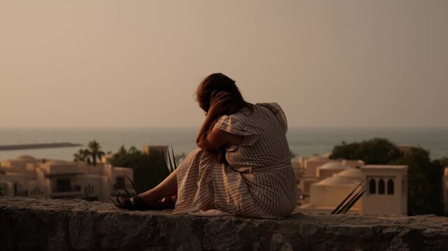 A woman in a striped dress sits on a stone wall overlooking the sea and traditional buildings. The warm sunset light creates a serene and reflective atmosphere. Woman relaxes on a stone ledge