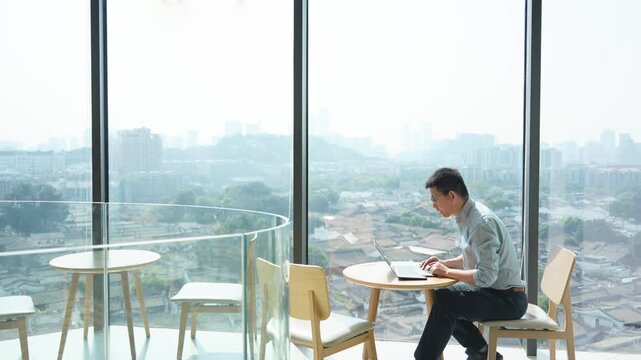 A young man working on a laptop by the window in a modern office with city view