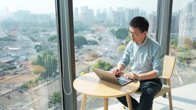 A young man working on a laptop by the window overlooking the cityscape