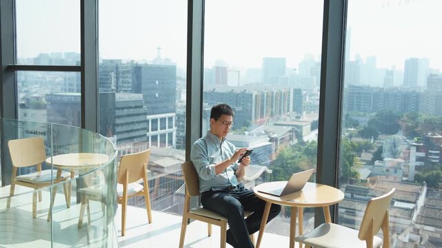 A young man working on a tablet computer at a modern caf&eacute; by the window with city skyline view