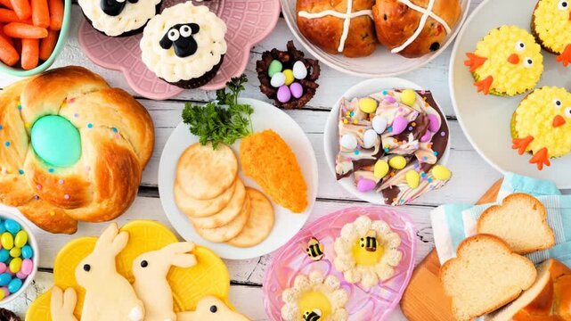 Easter table scene with a selection of breads, desserts and treats. Slow rotating zoom motion. Above view over a white wood background. Spring holiday food concept.