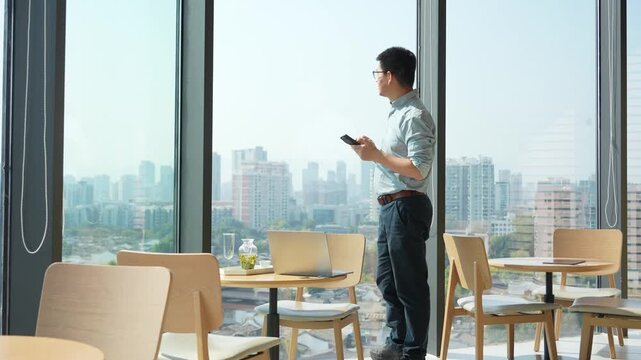 Young man standing by window in modern caf&eacute; holding smartphone while gazing at cityscape