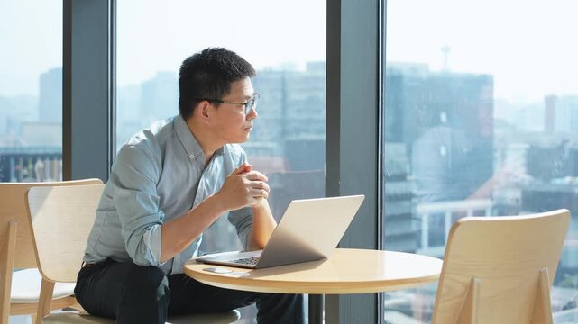 A young man wearing glasses works on a laptop by the window in a modern office with city view