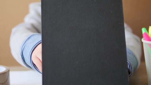 A close-up shot of a boy holding a black spiral-bound notebook upright, revealing the first page