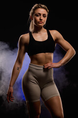 Fototapeta premium Confident athletic woman in black sports bra and shorts posing against dark smoky background exuding strength and empowerment