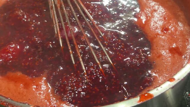 Close-up of a thick berry jam or compote cooking in a tall pot, with the mixture gently simmering and a whisk inside.