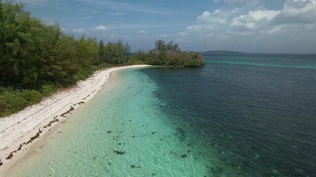 Aerial drone footage starting above a tropical beach, then flying over the island tip to reveal clear turquoise coastal waters and deep blue ocean under a bright, sunny sky.