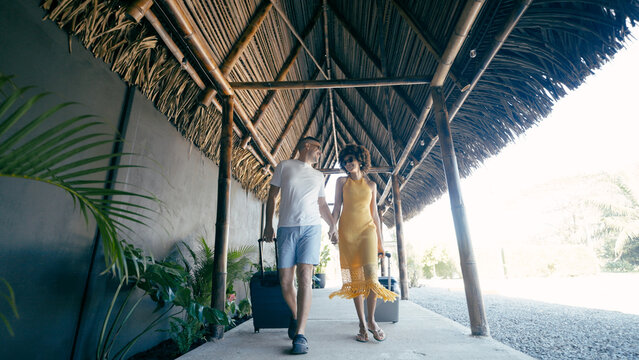 Multiracial couple holding hands, smiling while walking at a tropical resort, arriving for vacation with their bags