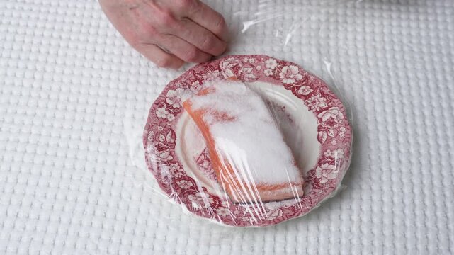 Wrapping salted salmon with plastic wrap. Hands wrapping a piece of salted salmon on a patterned plate with plastic wrap. Close-up food storage and preparation step on a white tablecloth.