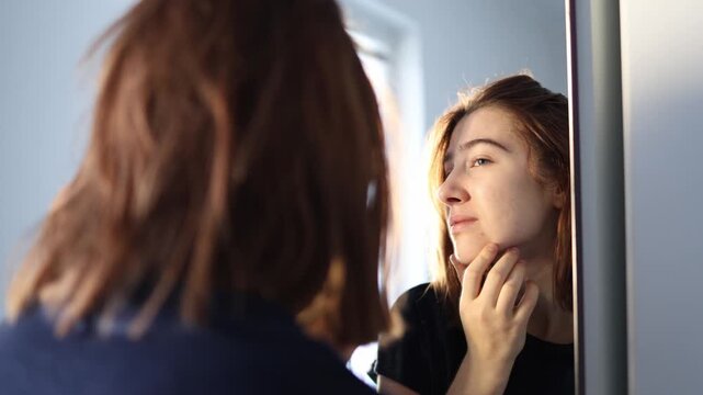 Young woman touches her chin looking at reflection with disappointment noticing skin imperfections morning self doubt frustration and insecurity