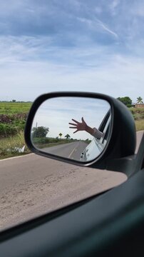 Video POV de viaje por carretera reflejado en el espejo lateral de un autom&oacute;vil, con una mano fuera de la ventana mientras se conduce por una carretera rural despejada bajo un cielo azul