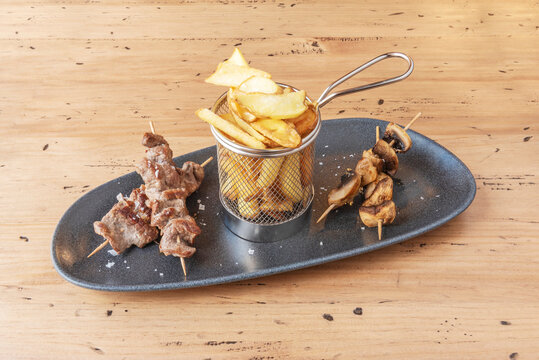 The perfect combination of smoky flavors and textures: marinated pork skewers with their characteristic caramelized sheen, served on a paper cone with thin fries and sprinkled