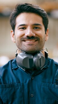 Portrait of a handsome young male carpenter with a mustache and hearing protection looking at the camera and smiling inside his workshop. Vertical
